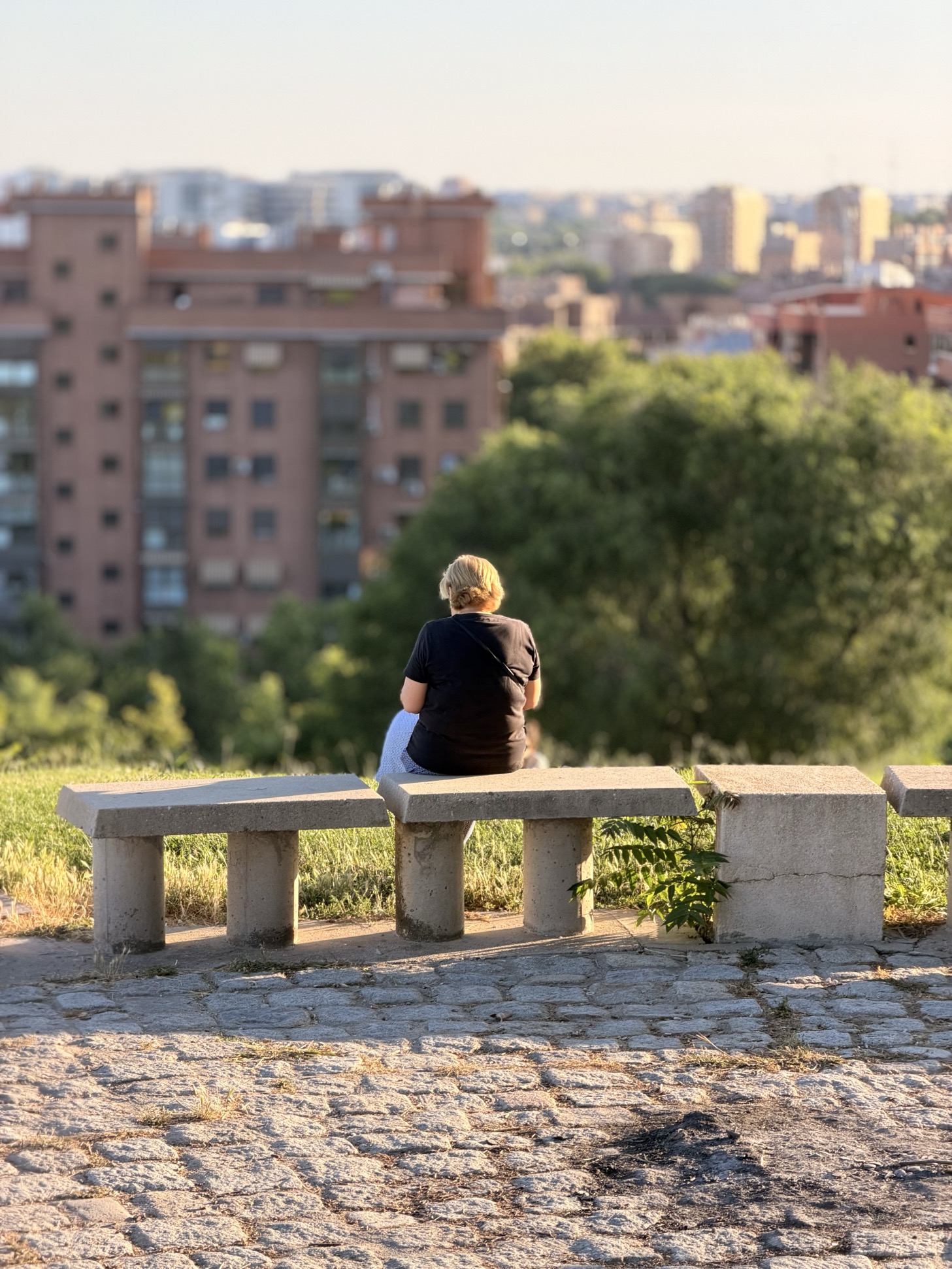 A person sits alone on a stone bench, seen from behind, looking out over a hazy Madrid skyline of apartment blocks and trees in warm late-afternoon light.