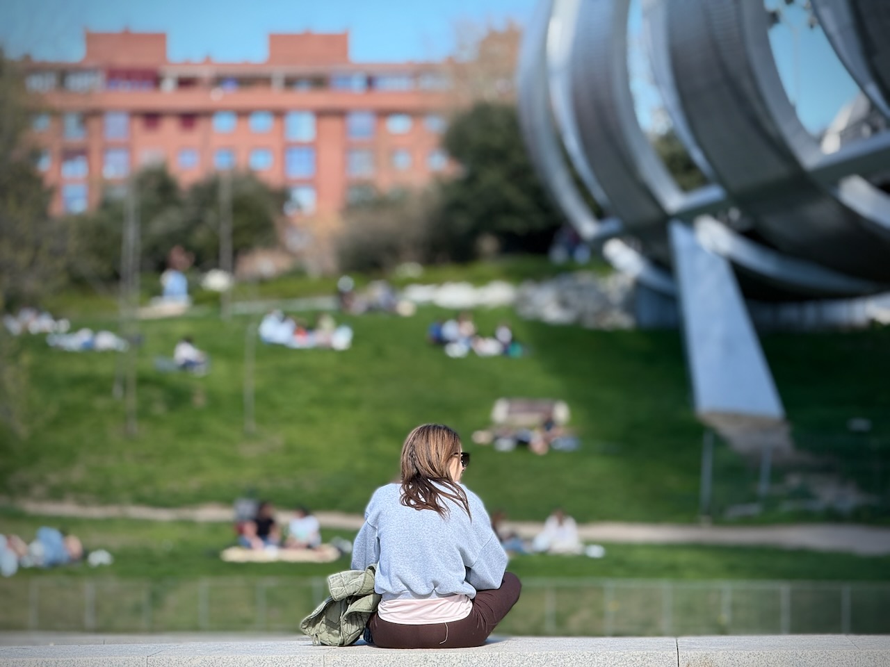 A woman sitting on a ledge overlooking a park with people relaxing on the grass.