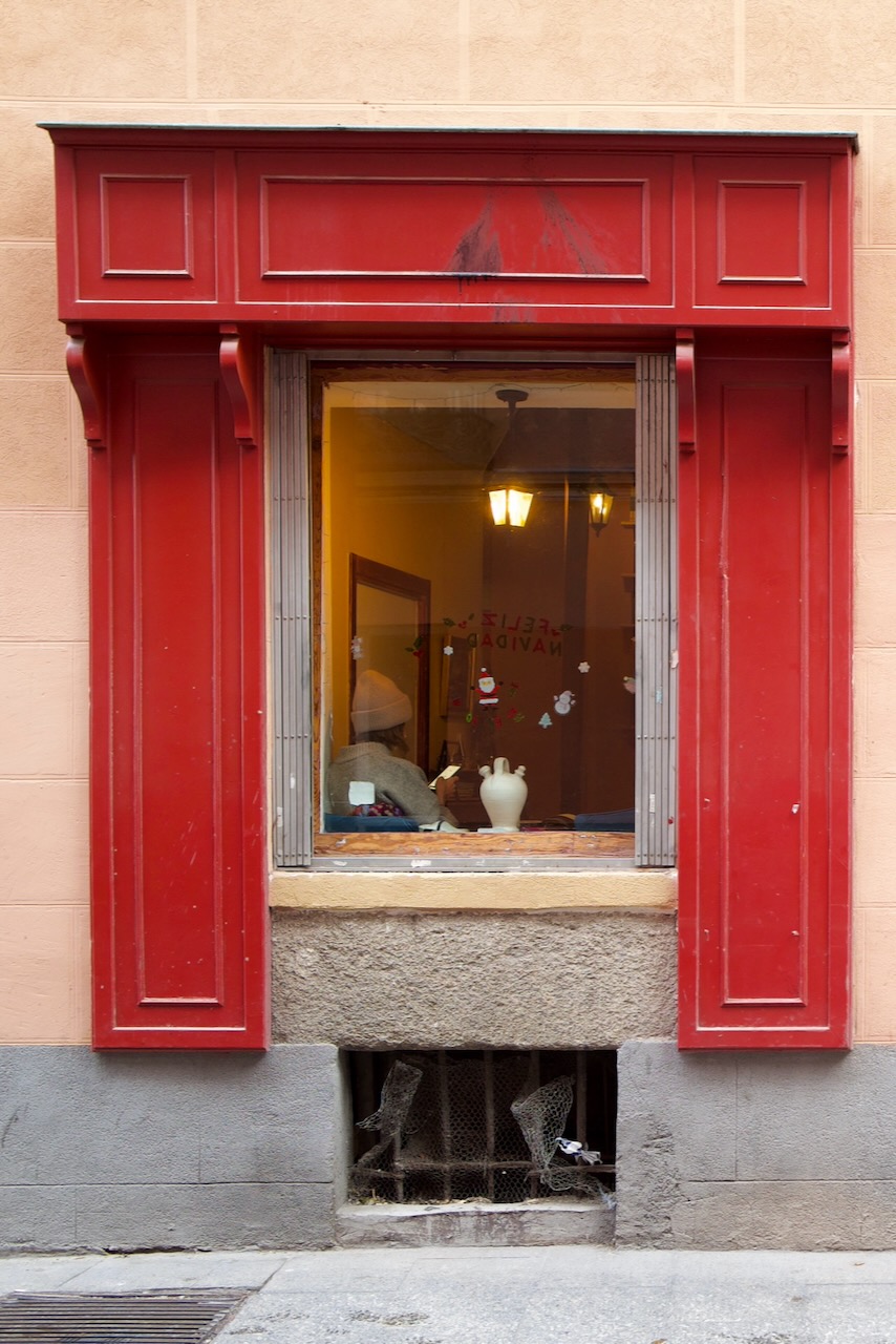 Red shutters frame a shop window with a lamp, vase, and reflections inside.