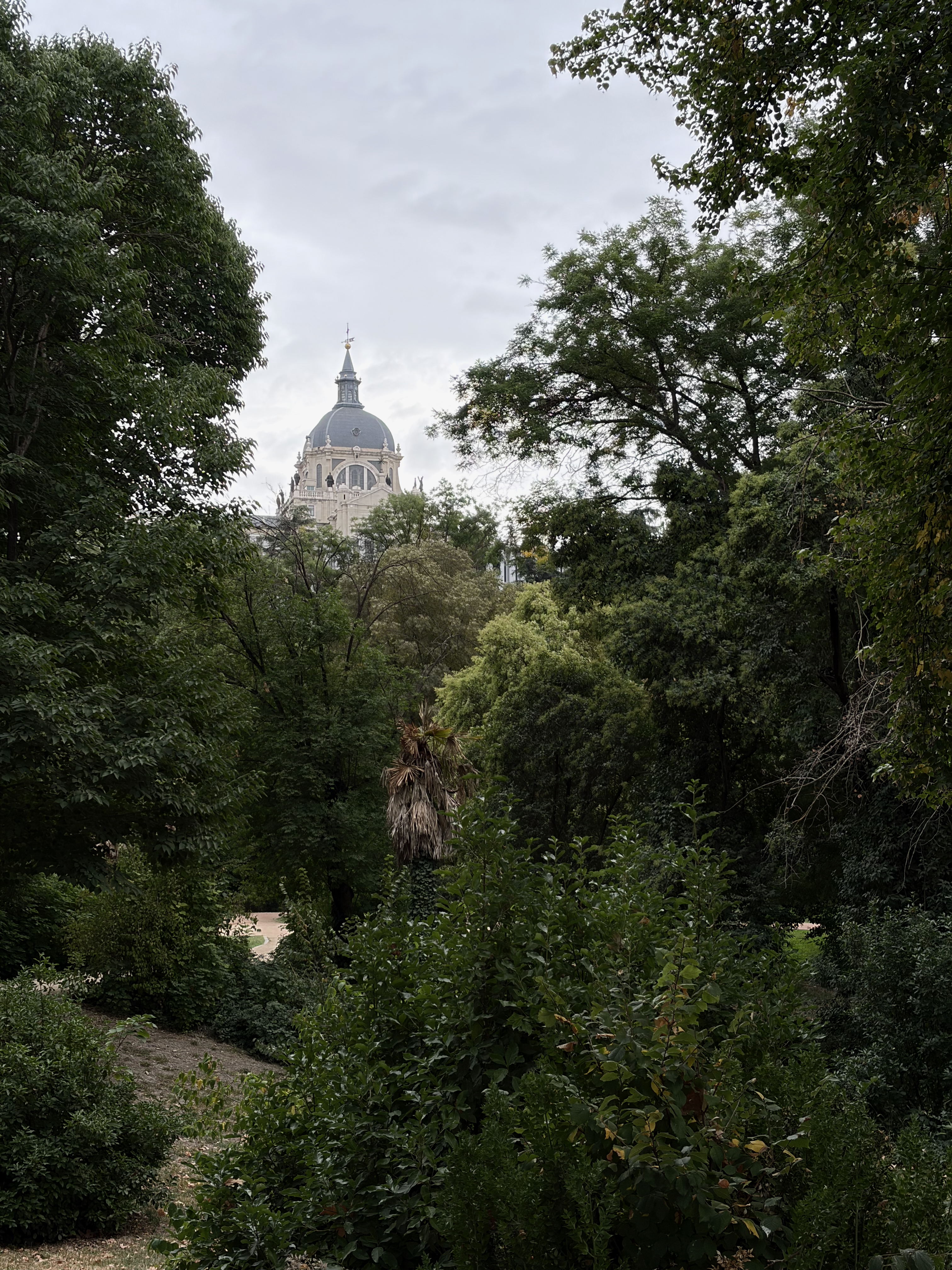 A domed building peeks through lush green trees under a cloudy sky.
